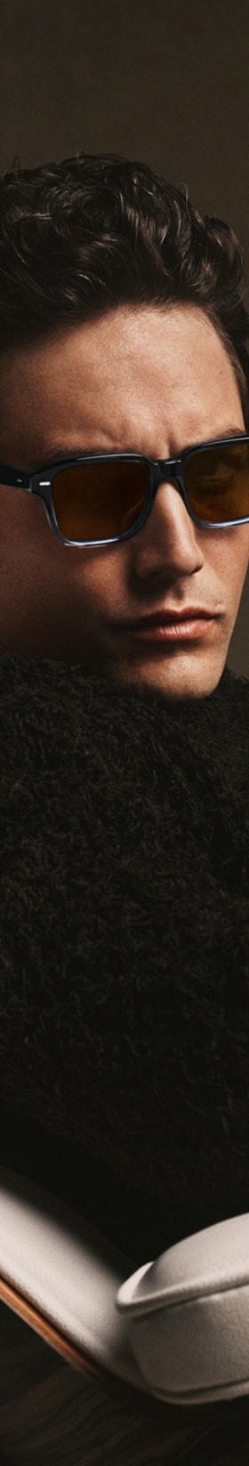 A man wearing tortoise Oliver Peoples sunglasses and a black shearling sweater, seated in a leather lounge chair
