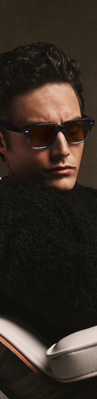 A man wearing tortoise Oliver Peoples sunglasses and a black shearling sweater, seated in a leather lounge chair