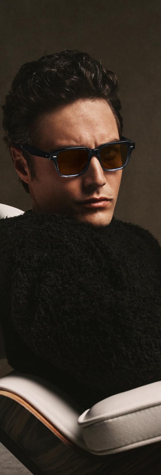 A man wearing tortoise Oliver Peoples sunglasses and a black shearling sweater, seated in a leather lounge chair