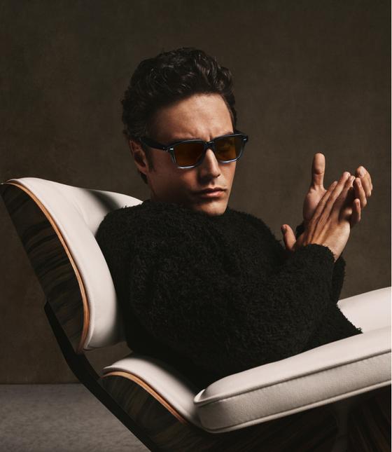 A man wearing tortoise Oliver Peoples sunglasses and a black shearling sweater, seated in a leather lounge chair