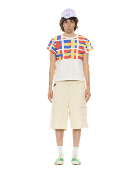 A man wearing a red, blue and yellow grid-print Organic Garmentz T-shirt with cream cargo shorts and a plaid cap