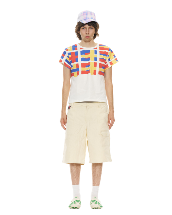 A man wearing a red, blue and yellow grid-print Organic Garmentz T-shirt with cream cargo shorts and a plaid cap