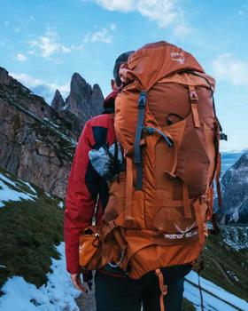 A hiker wearing a burnt-orange Osprey Aether 60 backpack on a snowy mountain ridge