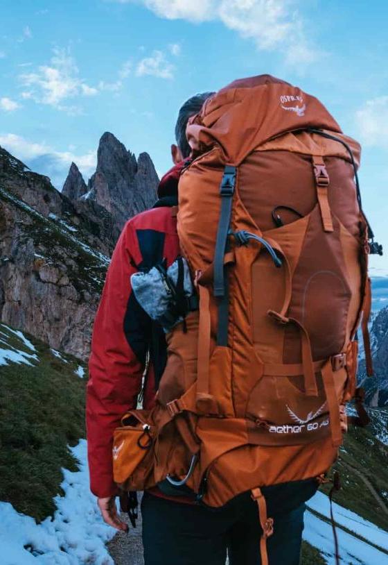 A hiker wearing a burnt-orange Osprey Aether 60 backpack on a snowy mountain ridge