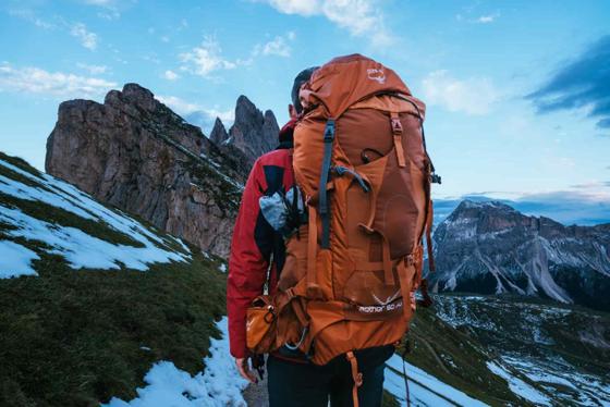 A hiker wearing a burnt-orange Osprey Aether 60 backpack on a snowy mountain ridge