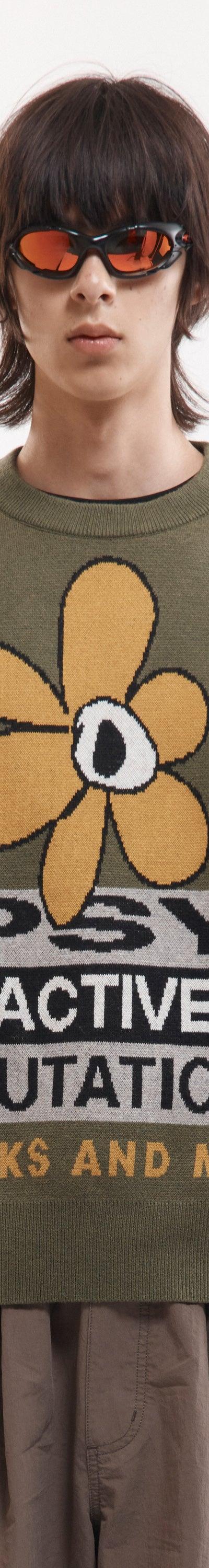 A man wearing an olive P.A.M. Perks and Mini intarsia knit sweater with a psychedelic flower motif