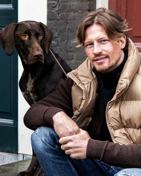 A man wearing a tan Panama Jack puffer vest over a brown knit sweater, crouching beside a dog