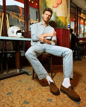 A man in a light denim shirt and washed jeans seated outside a Parisian café, wearing brown suede Paraboot derbies