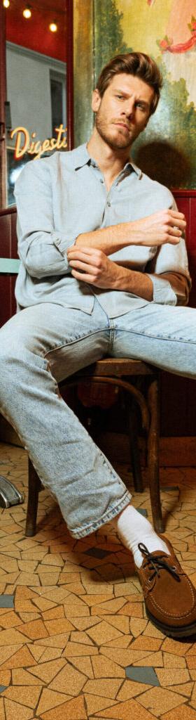 A man in a light denim shirt and washed jeans seated outside a Parisian café, wearing brown suede Paraboot derbies