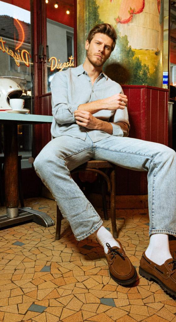 A man in a light denim shirt and washed jeans seated outside a Parisian café, wearing brown suede Paraboot derbies