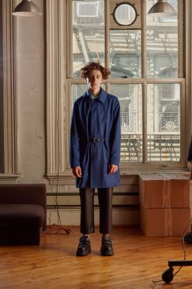A man wearing a blue belted Patrik Ervell mac coat with cropped black leather trousers