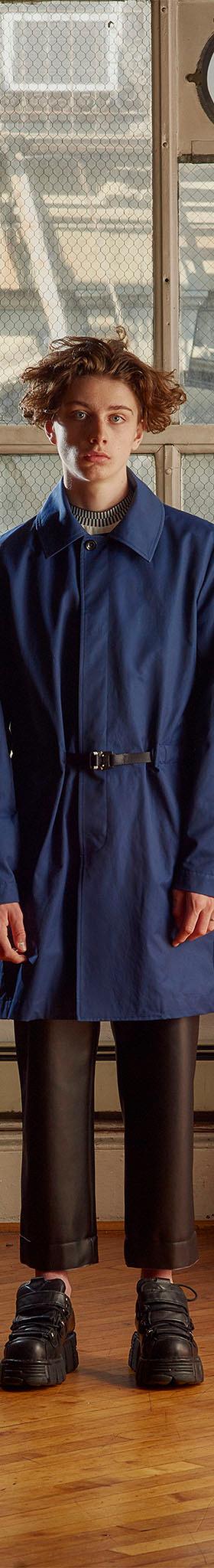 A man wearing a blue belted Patrik Ervell mac coat with cropped black leather trousers