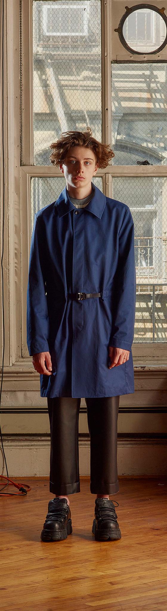 A man wearing a blue belted Patrik Ervell mac coat with cropped black leather trousers