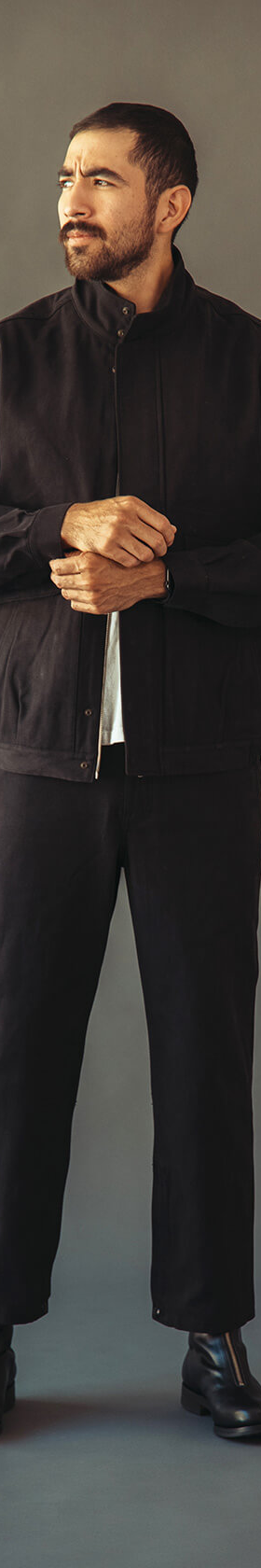 A man wearing a black Phigvel Makers & Co. work jacket with matching black trousers and leather boots