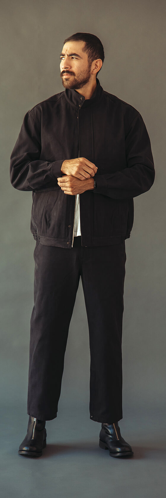 A man wearing a black Phigvel Makers & Co. work jacket with matching black trousers and leather boots