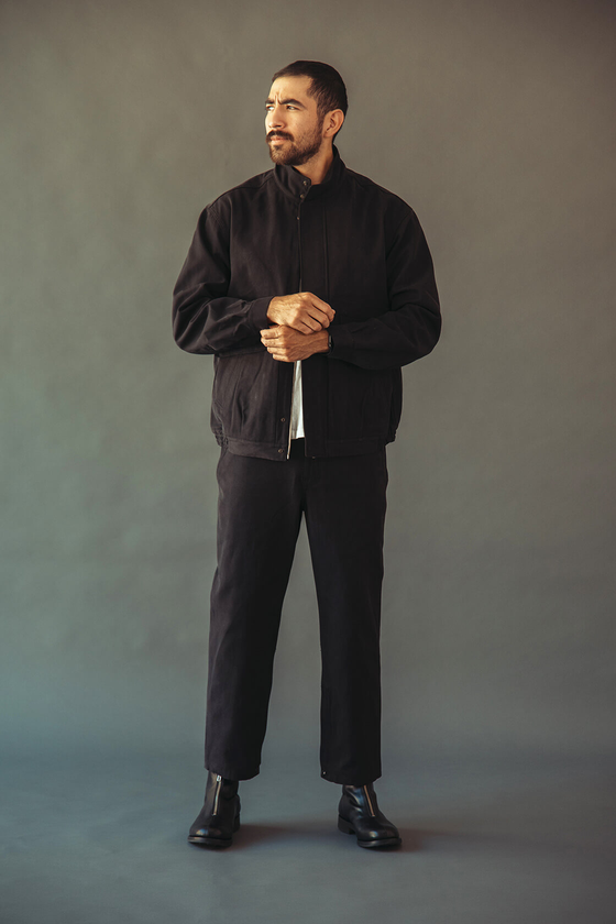 A man wearing a black Phigvel Makers & Co. work jacket with matching black trousers and leather boots