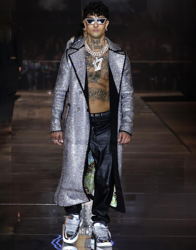 A man wearing a crystal-embellished silver Philipp Plein trench coat with leather trousers and metallic sneakers