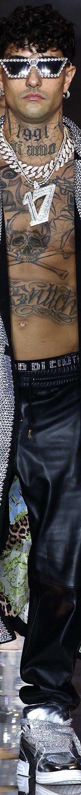 A man wearing a crystal-embellished silver Philipp Plein trench coat with leather trousers and metallic sneakers