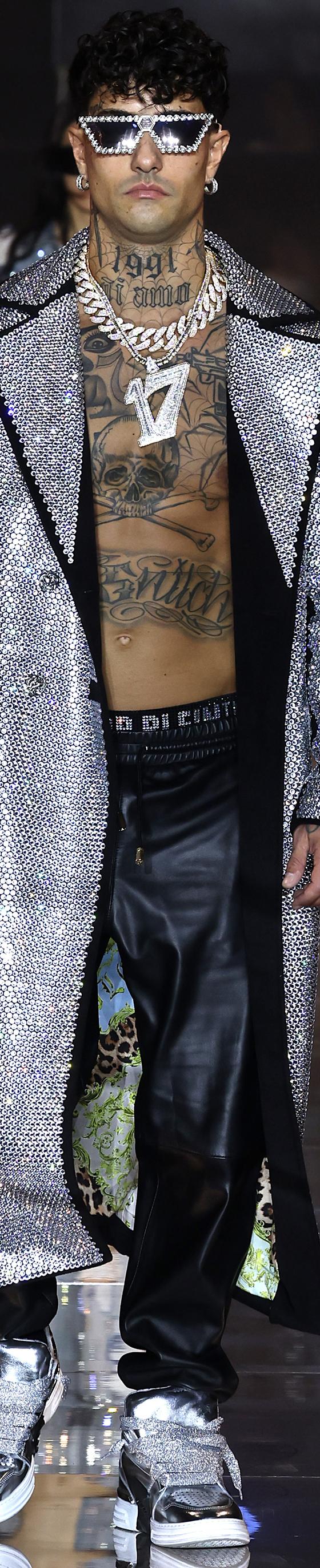 A man wearing a crystal-embellished silver Philipp Plein trench coat with leather trousers and metallic sneakers