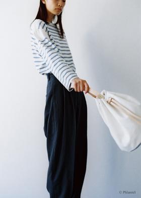 A woman wearing a blue and white striped Phlannèl top with dark wide-leg trousers, holding a white drawstring bag