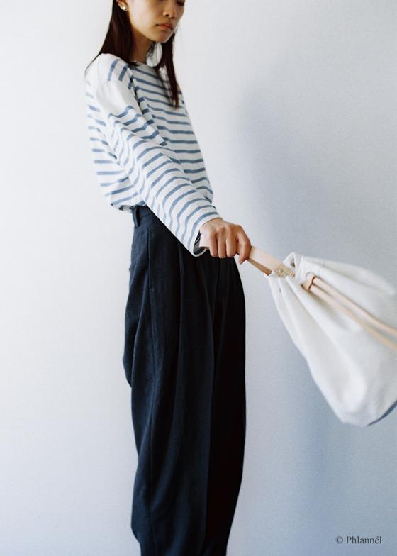 A woman wearing a blue and white striped Phlannèl top with dark wide-leg trousers, holding a white drawstring bag
