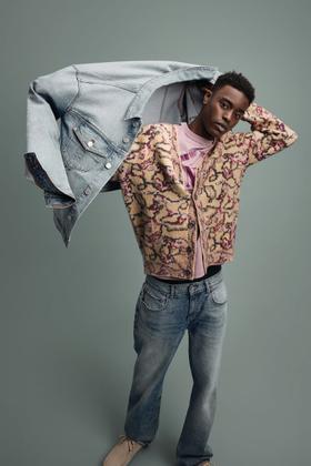 A man wearing a patterned fleece cardigan and faded jeans, holding a denim jacket overhead