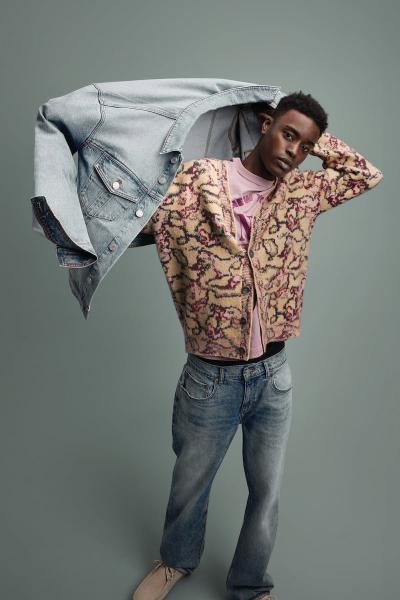A man wearing a patterned fleece cardigan and faded jeans, holding a denim jacket overhead
