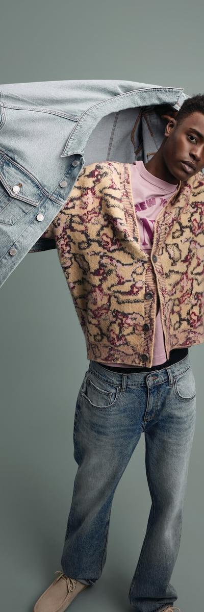 A man wearing a patterned fleece cardigan and faded jeans, holding a denim jacket overhead