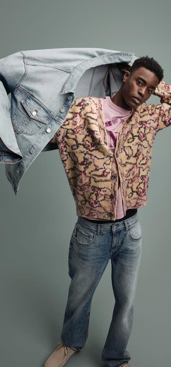 A man wearing a patterned fleece cardigan and faded jeans, holding a denim jacket overhead