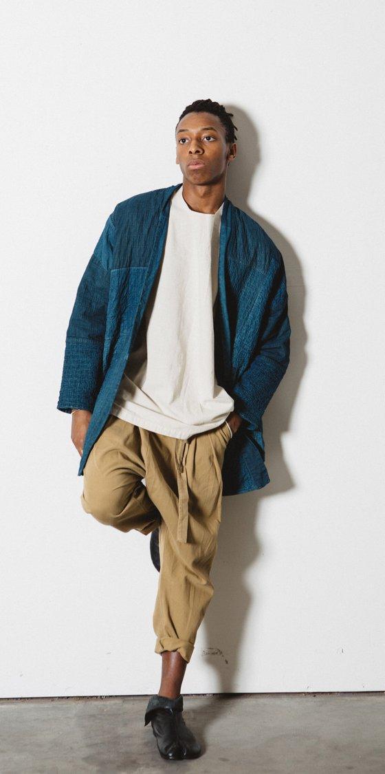 A man wearing an indigo quilted Prospective Flow haori jacket over a cream tee and tan cropped trousers
