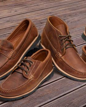 A group of brown Quoddy handsewn leather moccasins, a penny loafer, a chukka and a boat shoe arranged on a wooden deck