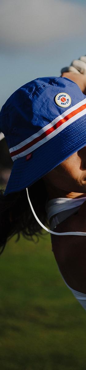 A woman wearing a blue and red striped Random Golf Club bucket hat mid golf swing on a course