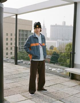 A man wearing a navy and orange Rassvet zip jacket, brown wide-leg corduroy trousers and a black beanie on a Moscow rooftop