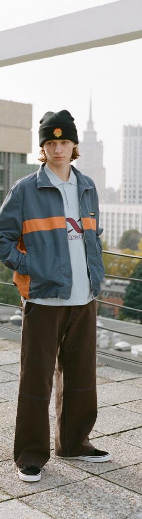 A man wearing a navy and orange Rassvet zip jacket, brown wide-leg corduroy trousers and a black beanie on a Moscow rooftop