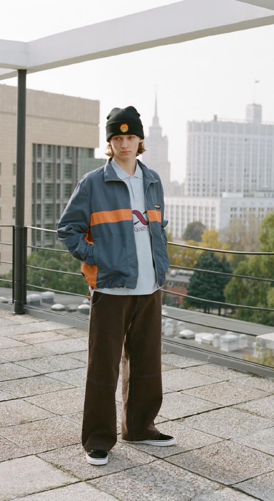A man wearing a navy and orange Rassvet zip jacket, brown wide-leg corduroy trousers and a black beanie on a Moscow rooftop