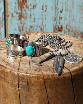 Hand-stamped sterling silver rings with turquoise stones and a feather pendant necklace arranged on a weathered wood stump