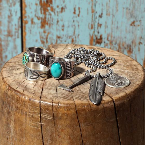 Hand-stamped sterling silver rings with turquoise stones and a feather pendant necklace arranged on a weathered wood stump