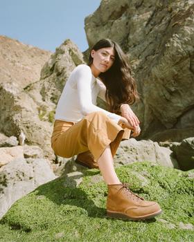 A woman wearing tan Red Wing moc toe boots with camel trousers and a white long-sleeve top, crouched on a mossy coastal rock