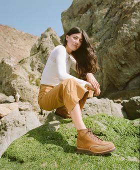 A woman wearing tan Red Wing moc toe boots with camel trousers and a white long-sleeve top, crouched on a mossy coastal rock