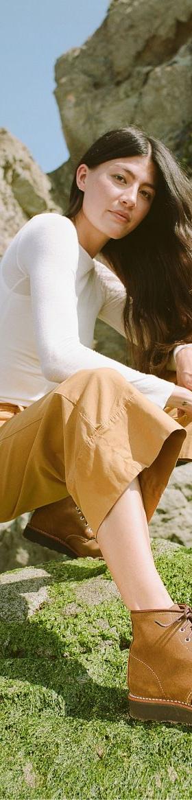 A woman wearing tan Red Wing moc toe boots with camel trousers and a white long-sleeve top, crouched on a mossy coastal rock