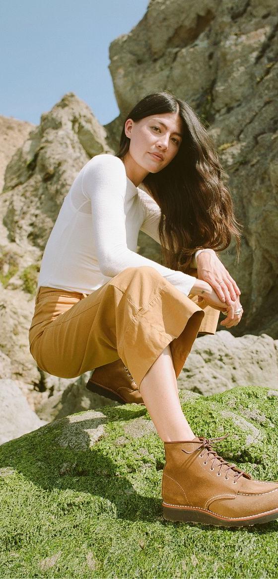 A woman wearing tan Red Wing moc toe boots with camel trousers and a white long-sleeve top, crouched on a mossy coastal rock