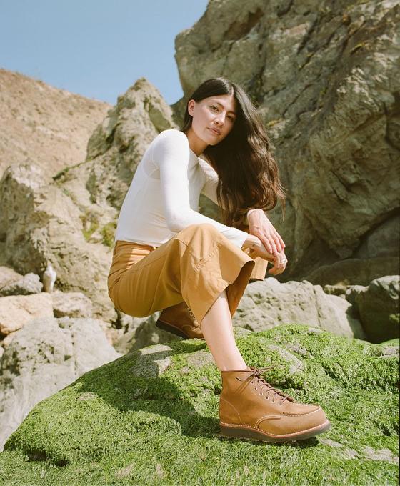 A woman wearing tan Red Wing moc toe boots with camel trousers and a white long-sleeve top, crouched on a mossy coastal rock