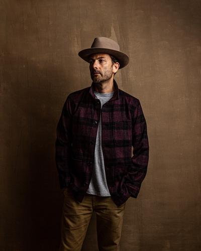 A man wearing a burgundy plaid wool overshirt with a grey tee, tan trousers and a felt hat