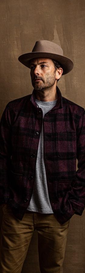 A man wearing a burgundy plaid wool overshirt with a grey tee, tan trousers and a felt hat