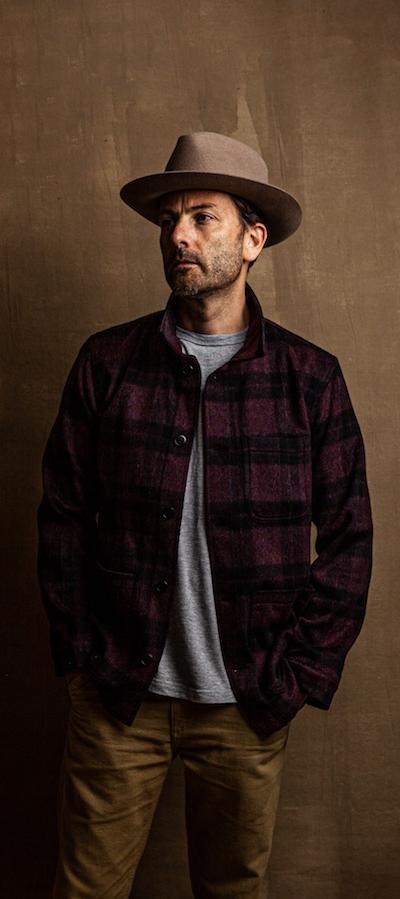 A man wearing a burgundy plaid wool overshirt with a grey tee, tan trousers and a felt hat
