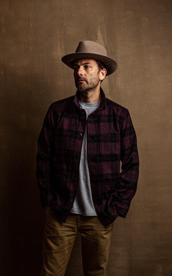 A man wearing a burgundy plaid wool overshirt with a grey tee, tan trousers and a felt hat