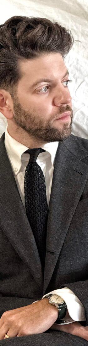 A man wearing a charcoal Rochester Tailored Clothing suit jacket with a white shirt and knit tie