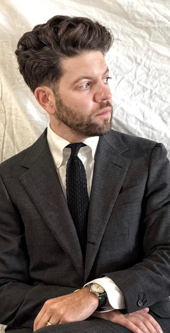 A man wearing a charcoal Rochester Tailored Clothing suit jacket with a white shirt and knit tie