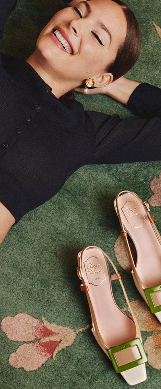 A smiling woman in a black cropped cardigan and pink skirt lying on a green floral carpet beside green and white Roger Vivier buckled slingback pumps