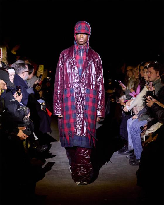 A man wearing a glossy burgundy Rolf Ekroth puffer coat over a red tartan check ensemble with a matching flat cap
